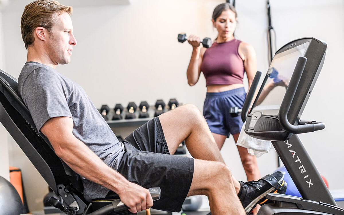 Man riding a Matrix recumbent bike with woman working out in the background